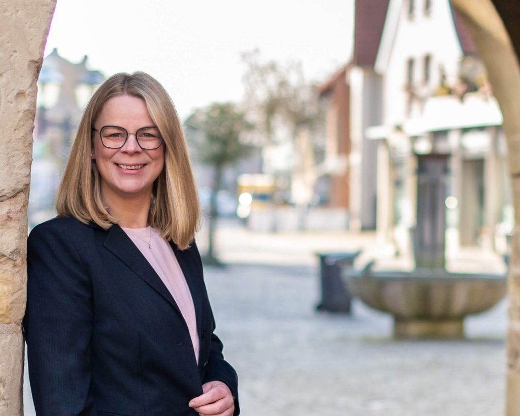 Geraldine am Torbogen Geraldine Henneböhl, Bürgermeisterkandidatin für Havixbeck und Hohenholte, steht am Torbogen. Sie lächelt freundlich und strahlt Unabhängigkeit aus. Im Hintergrund ist der Brunnen zu sehen.